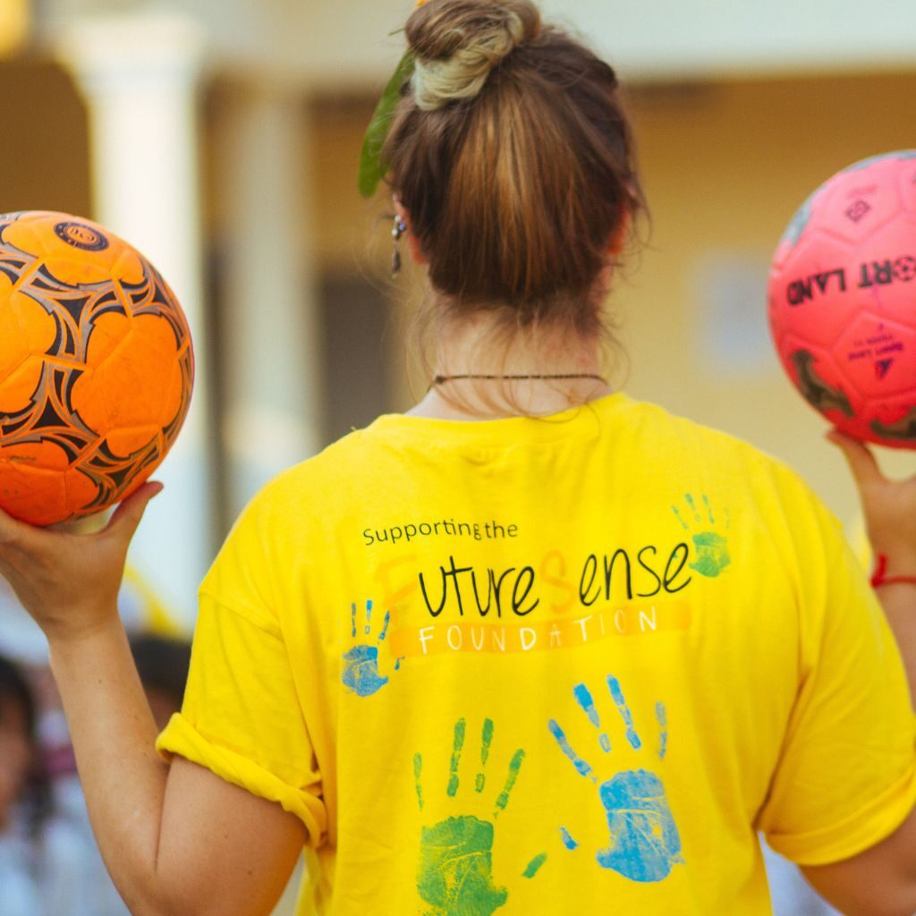A young woman facing away from the camera holding two footballs and wearing a yellow FutereSense Foundation t-shirt - GapGuru
