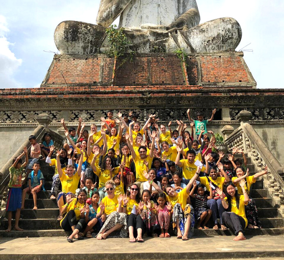 A large group of gap year students and local Cambodian children sat on the steps of a large statue of Buddha waving their arms and smiling - GapGuru