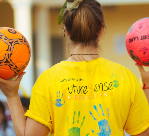 A young woman facing away from the camera holding two footballs and wearing a yellow FutereSense Foundation t-shirt - GapGuru